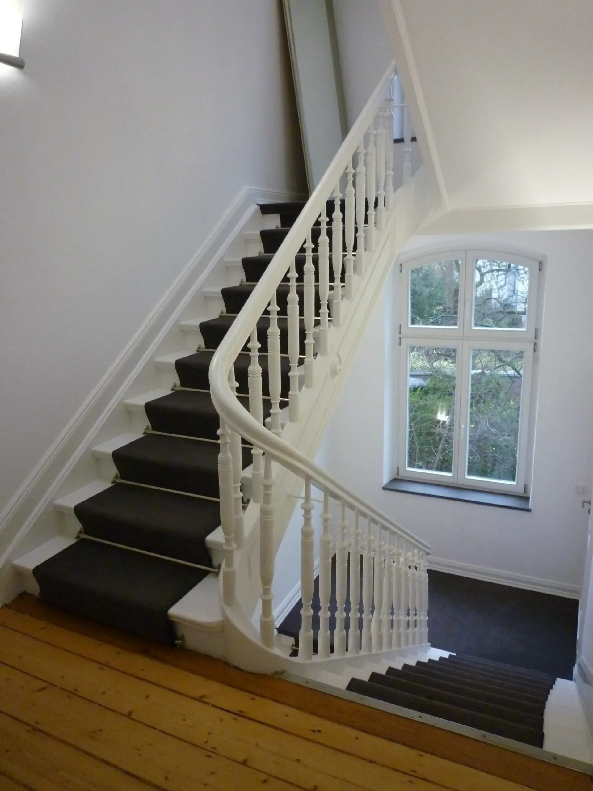 Ansicht einer frisch renovierten, klassischen Wendeltreppe mit elegantem, weiß lackiertem Geländer und dunklem Teppichläufer.