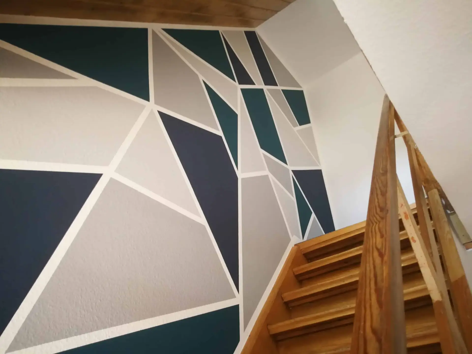 Kreative Wandgestaltung in einem Treppenhaus mit einem großflächigen, geometrischen Muster aus Dreiecken in Blau-, Grün- und Grautönen.